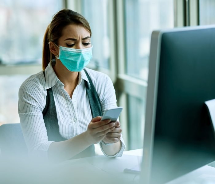 young-businesswoman-with-face-mask-texting-smart-phone-while-working-office